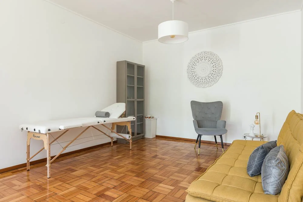 Sala de estética com maca, cadeira cinza, sofá amarelo, parede branca, piso de madeira, luminária branca e decoração.