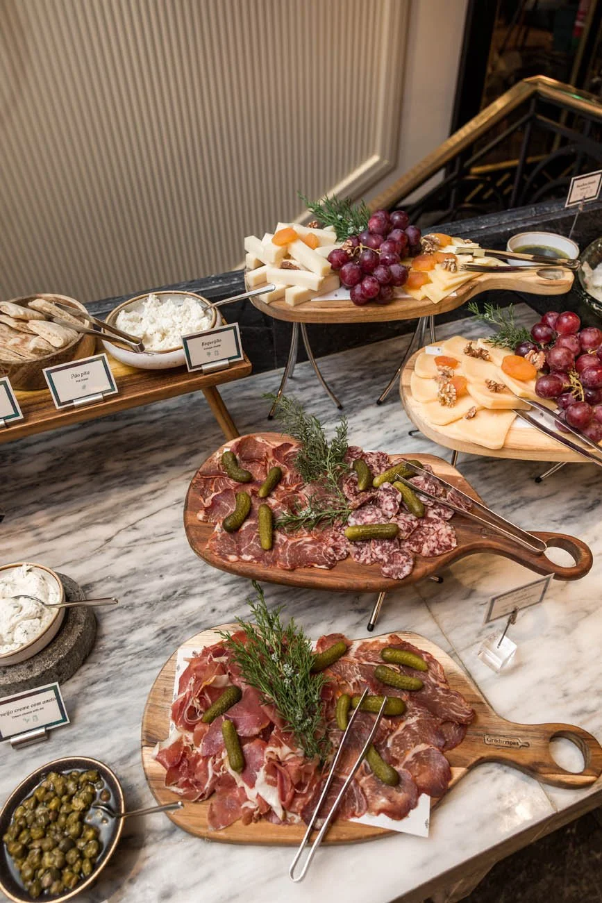 Mesa de frios com vários tipos de queijos, uvas, presunto, salame, pepinos, creme e picles, decorada com folhagens verdes.