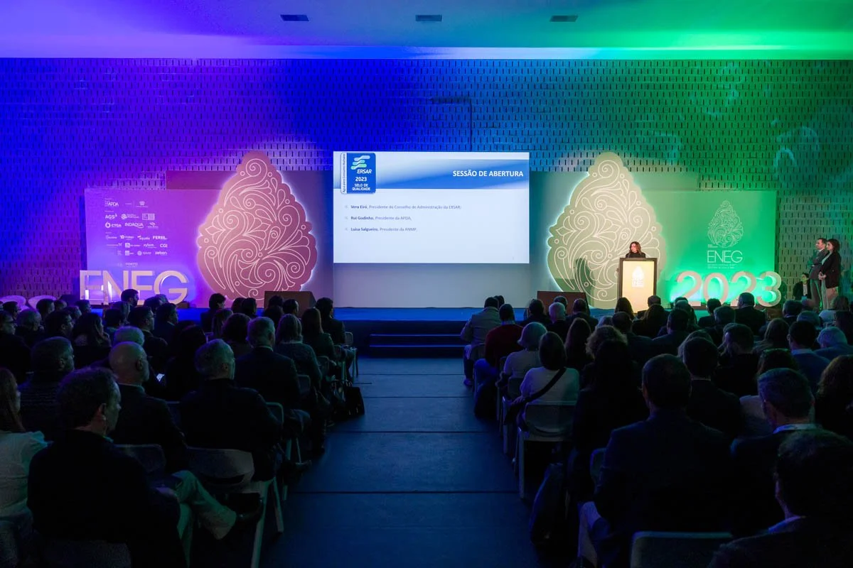 Audiência em uma conferência com pessoas sentadas assistindo a uma apresentação no palco. O palco tem uma grande tela de projeção, com uma frase em português e símbolos no fundo, decorado com elementos gráficos iluminados em tons de azul, verde e rox