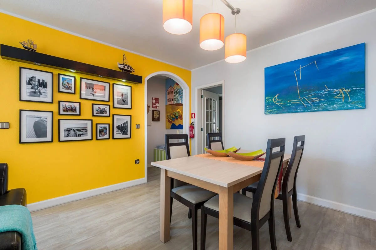 Sala de jantar com mesa e quatro cadeiras, parede amarela com fotos emolduradas, parede branca com pintura azul e luminárias laranja no teto.