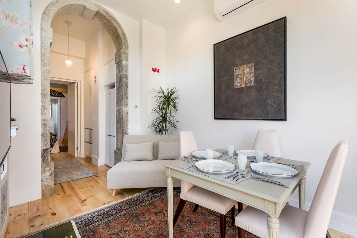 Sala de jantar com mesa branca, pratos e copos, cadeiras brancas e quadro preto na parede.