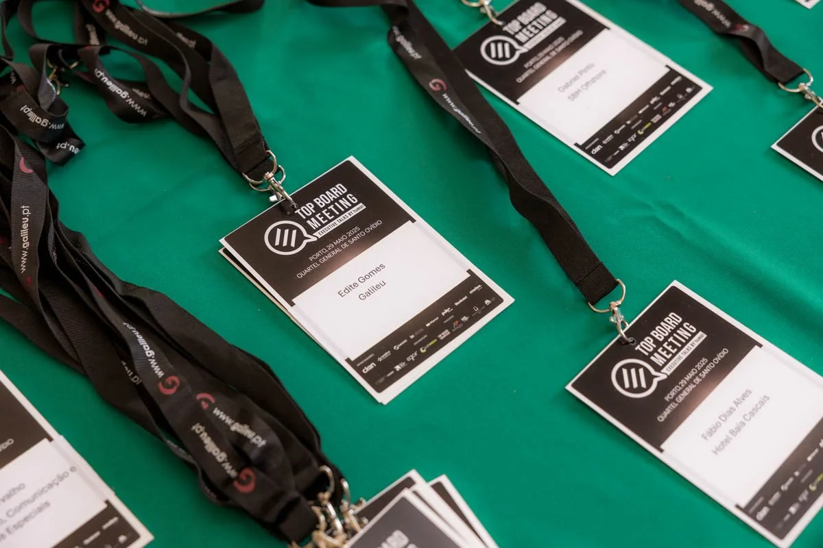 Credenciais de participante de uma conferência intitulada "Top Board Meeting" com nomes como Edite Gomes Gauleu e Fábio Ovas Alves, colocadas sobre uma mesa verde.