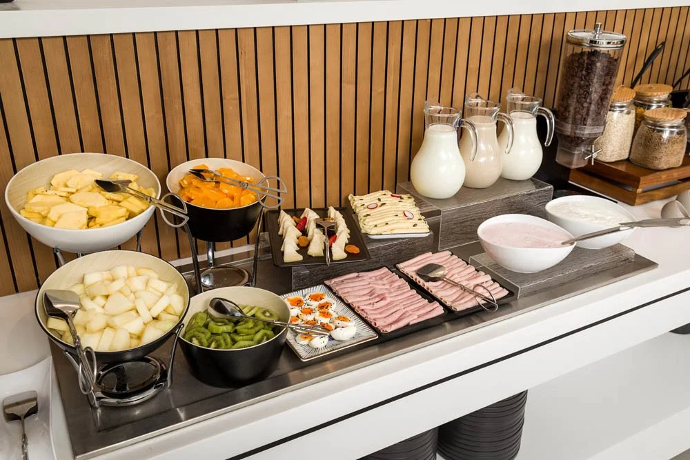 Estação de café da manhã com frutas, iogurte, ovos, presunto, pratos com queijo e pão, além de cereais e leite.