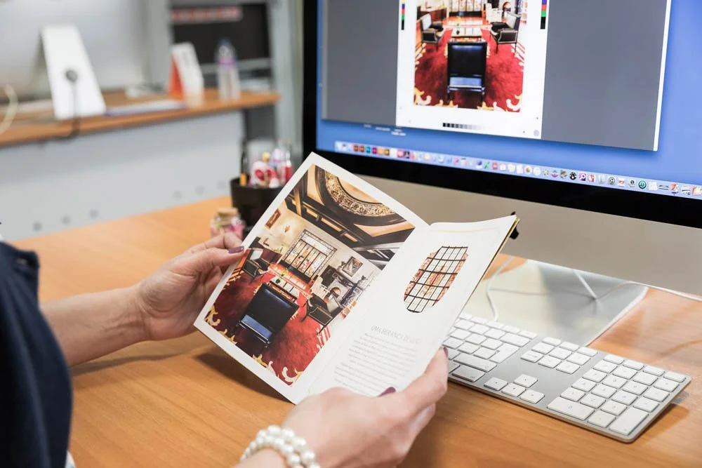 Pessoa segurando um catálogo ou revista com fotos de interiores de uma sala de reuniões ou escritório moderno, com uma mesa de reuniões, cadeiras e uma iluminação elegante, ao lado de um computador com uma tela de design de interiores