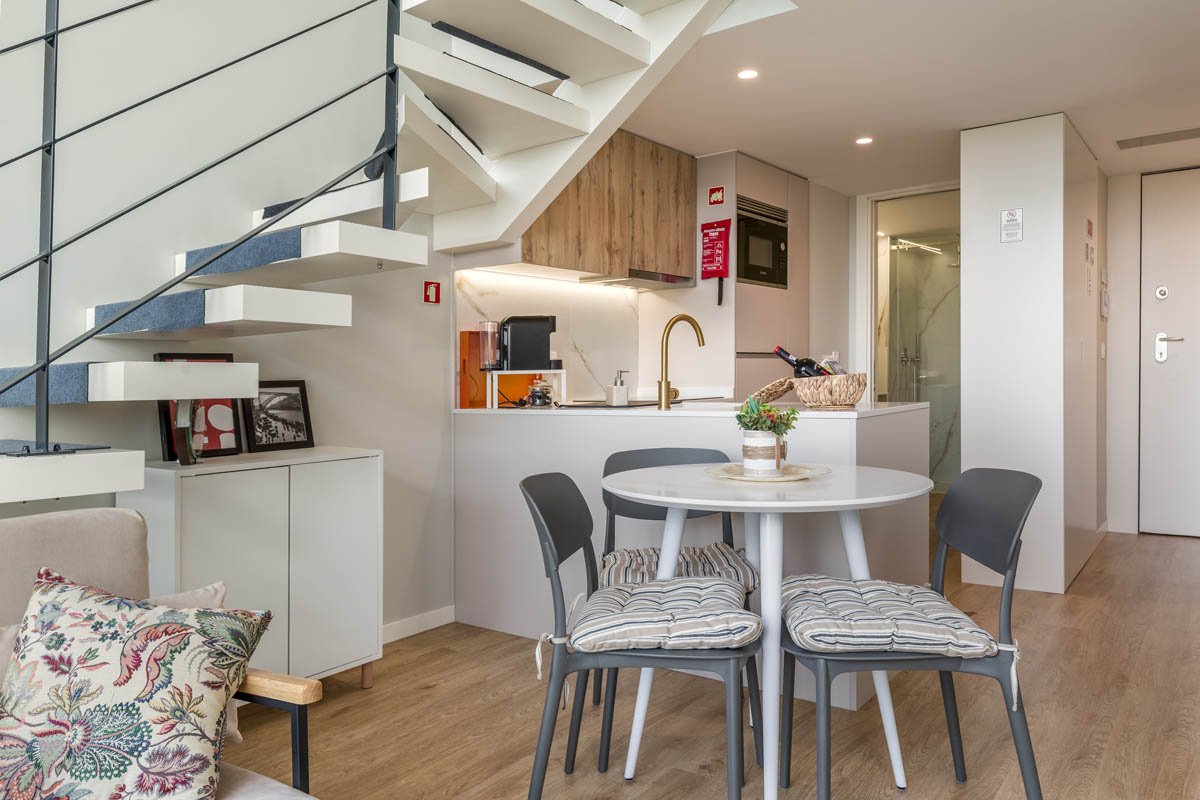 Interior de uma sala de jantar e cozinha compacta com escada de metal e madeira, mesa redonda com quatro cadeiras com almofadas, e detalhes decorativos.