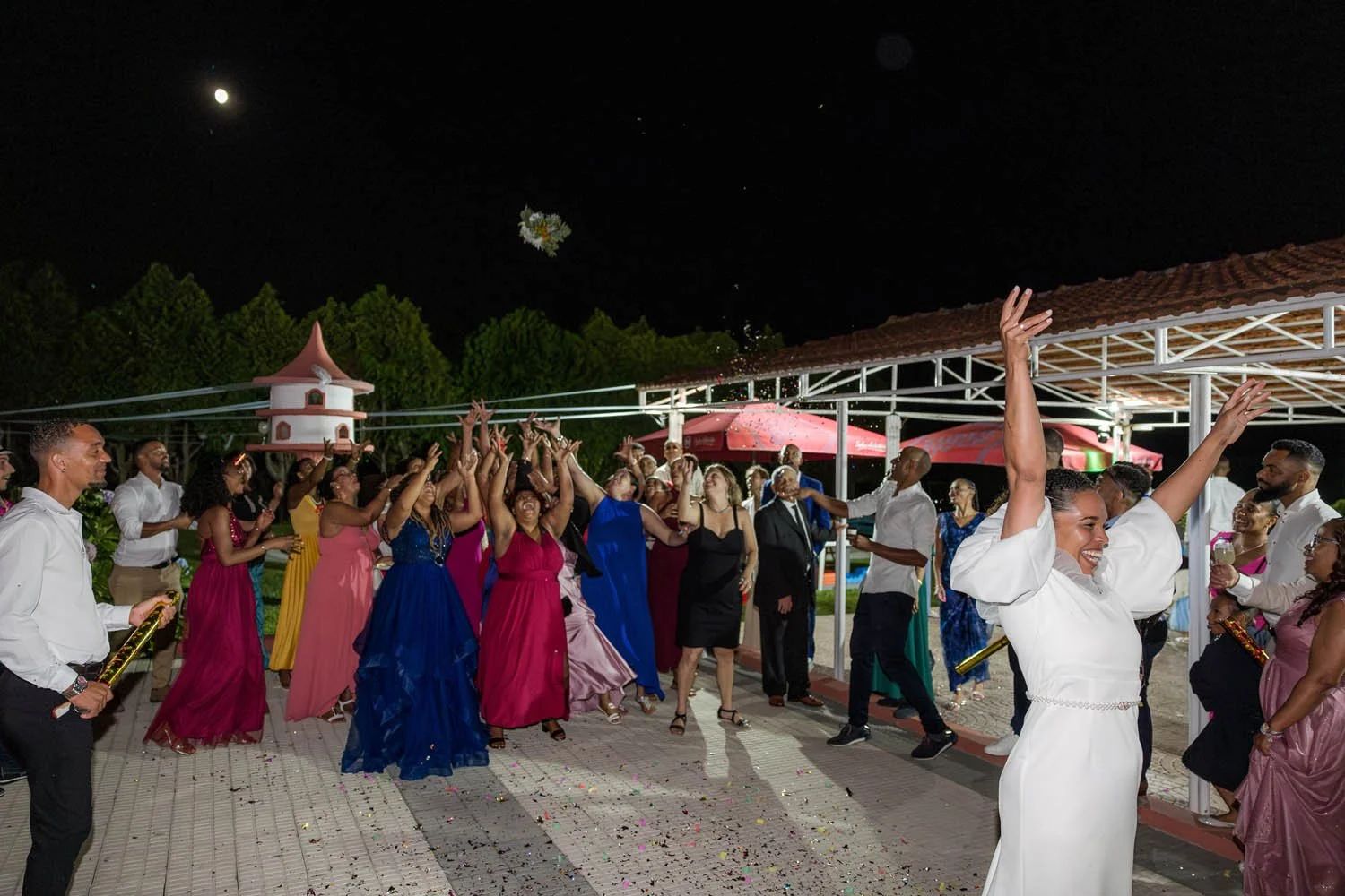 Pessoas celebrando ao ar livre à noite, jogando flores no ar, com uma lua visível no céu, em uma festa com decoração e música.