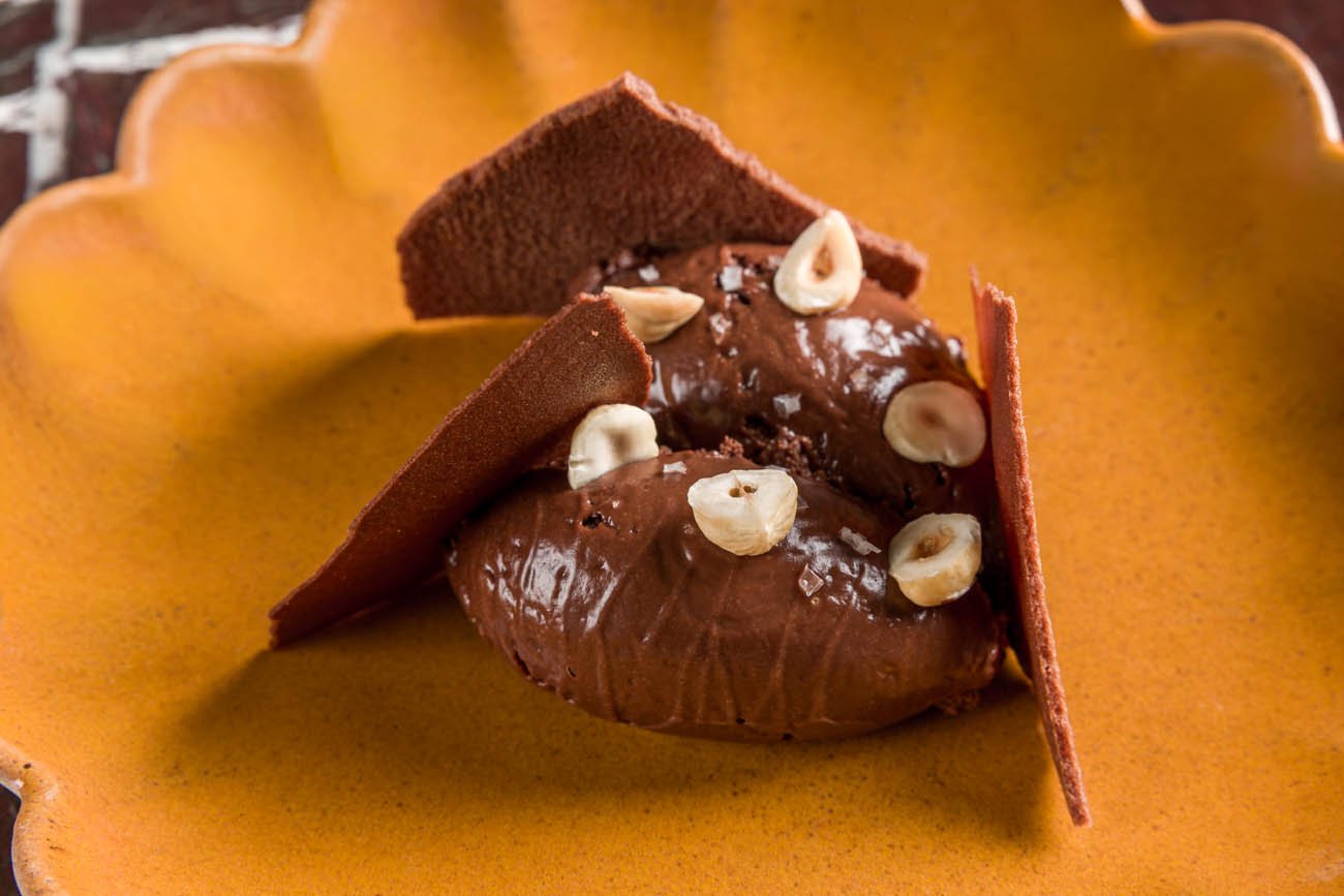 Sobremesa de chocolate servida em um prato de cerâmica laranja, com duas porções cobertas de chocolate, decoradas com castanhas e biscoitos de chocolate.