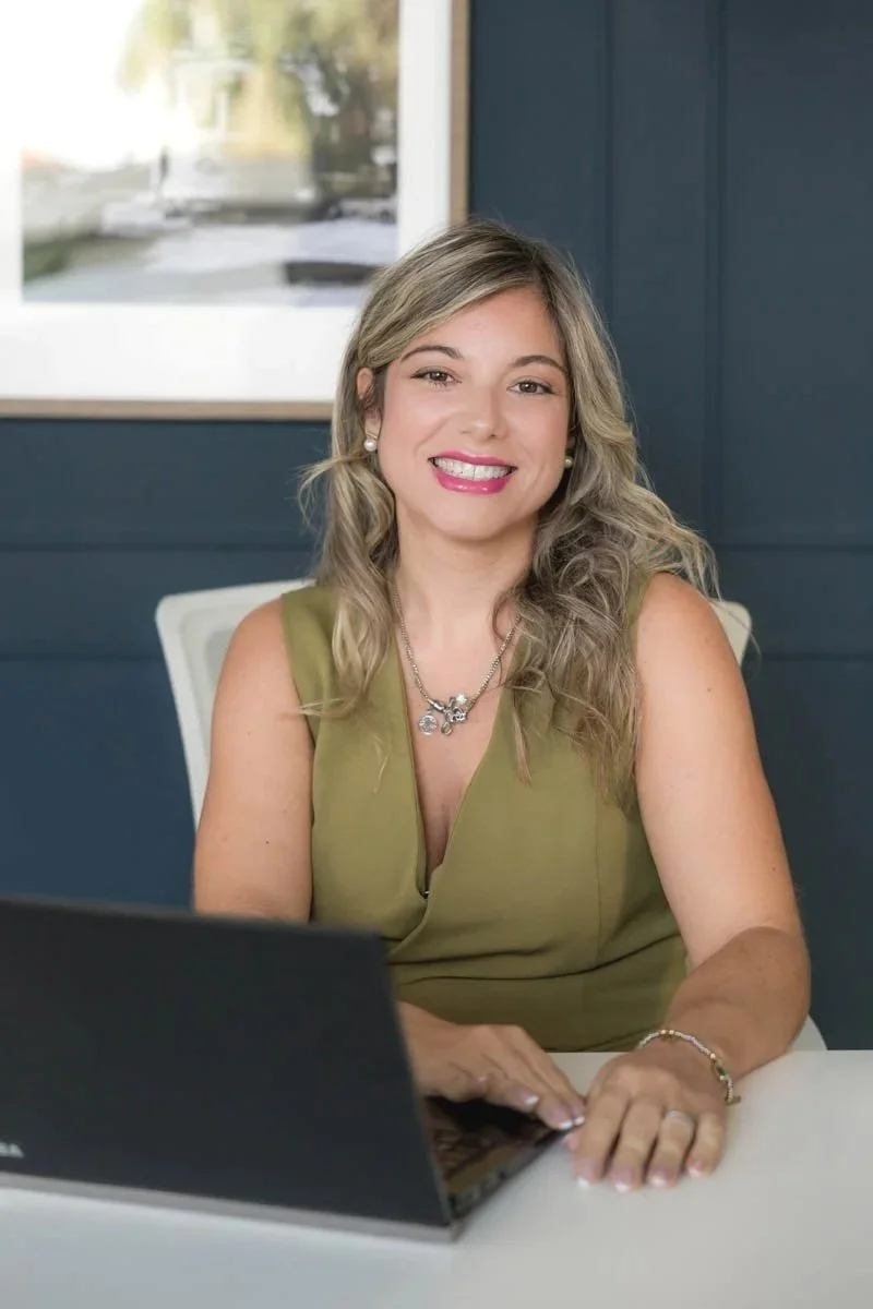 Mulher sorridente com cabelo loiro ondulado, usando vestido verde, brincos de pérola, colar e pulseira, sentada em uma mesa ao lado de um laptop, em um escritório com parede azul e janela ao fundo.