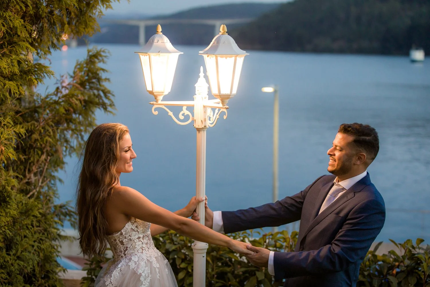 Casal de noivos sorridentes segurando uma luminária de rua ao pôr do sol na beira de um lago com árvores ao fundo.