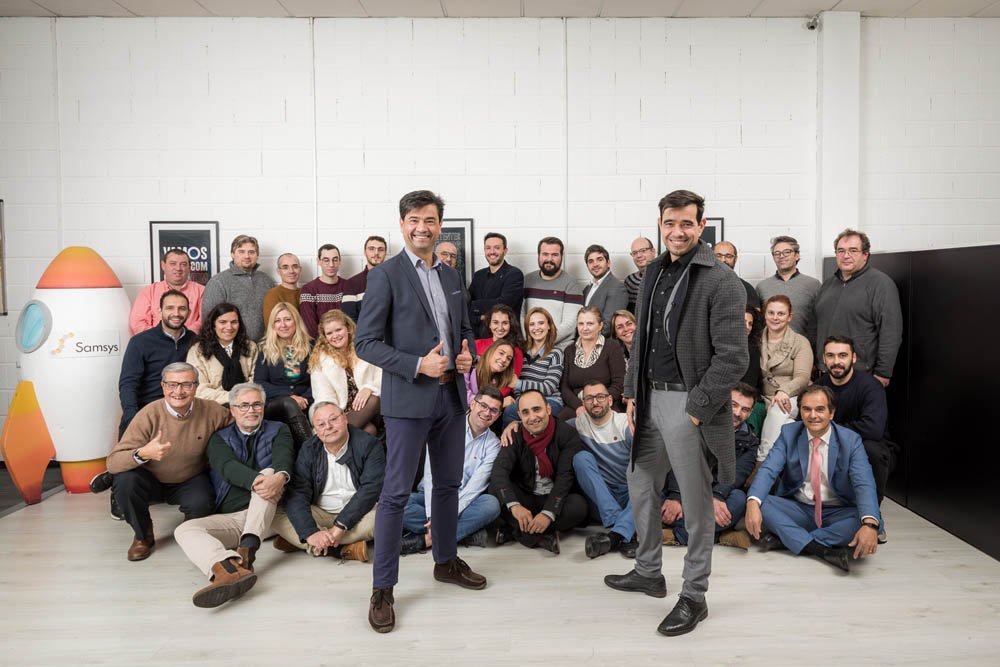 Grupo de pessoas posando em uma sala com decoração moderna, incluindo uma nave espacial de brinquedo e quadros nas paredes, com dois homens de terno na frente, sorrindo.