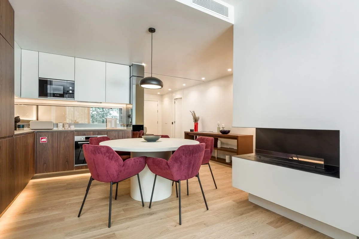 Sala de jantar com mesa redonda branca, cadeiras de veludo rosa, área de cozinha moderna com armários brancos e madeira escura, lareira embutida na parede branca, decoração com vaso de flores vermelhas e pratos.