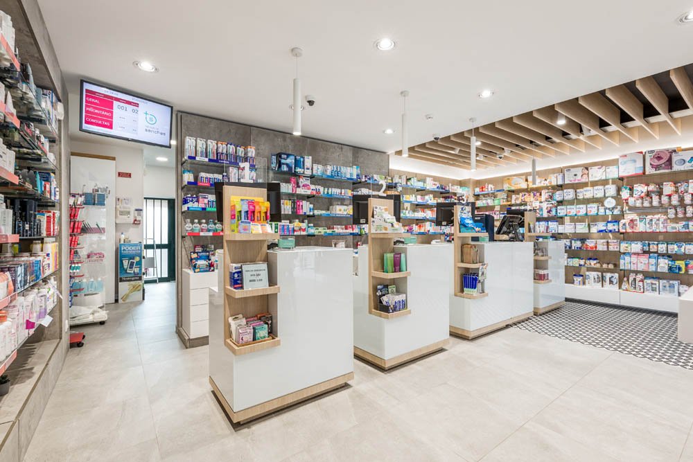 Interior de uma farmácia com balcões, prateleiras com medicamentos e produtos de higiene, e telas de computador na bancada.