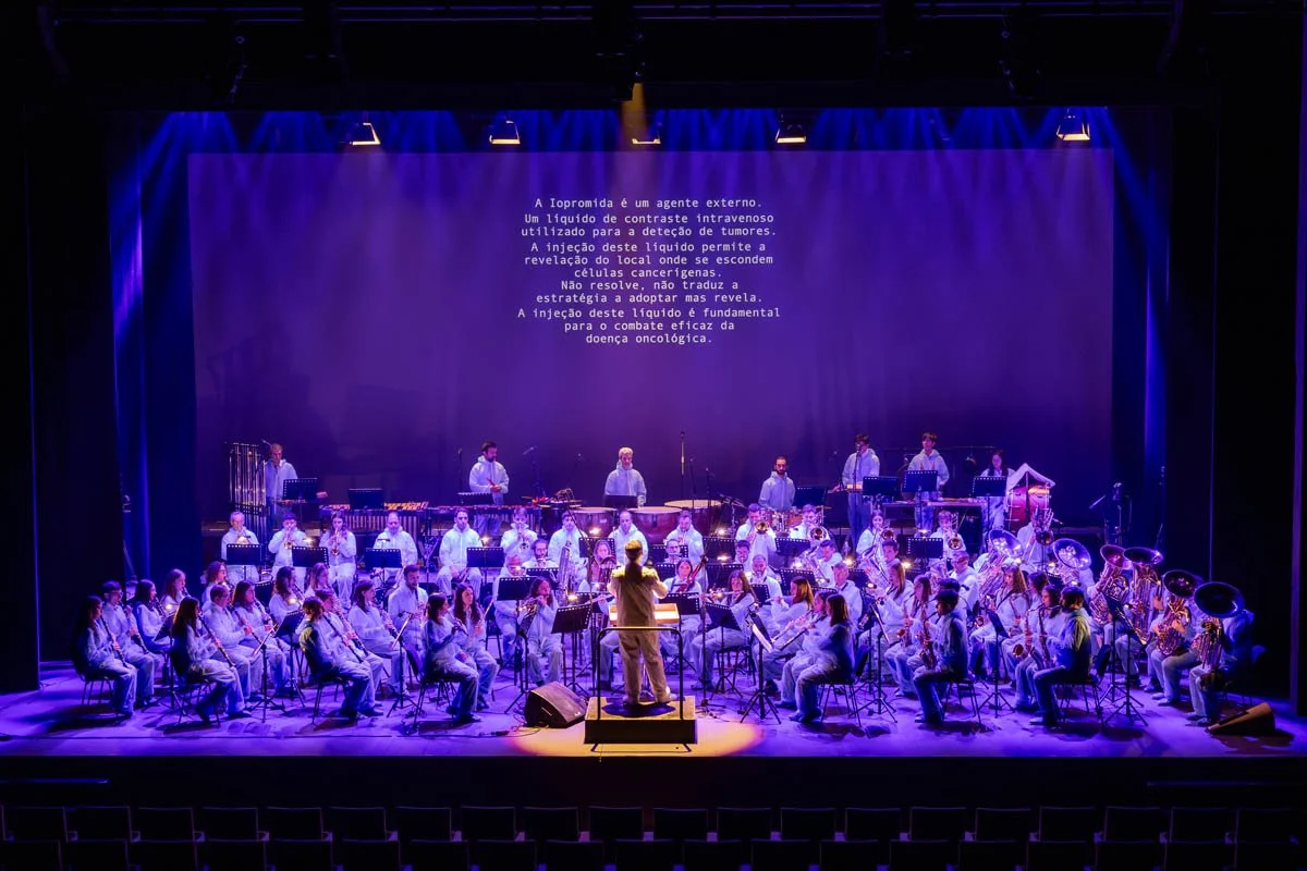 Orquestra tocando em palco com iluminação azul, com uma pessoa conduzindo na frente, e uma tela com texto em português acima.