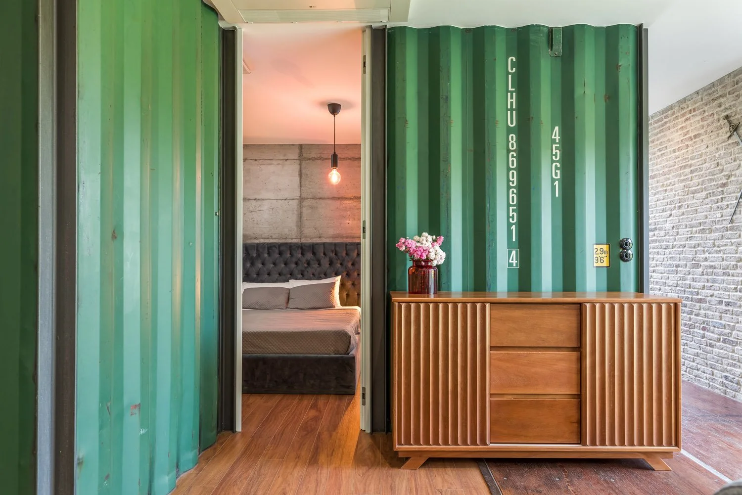 Entrada de uma sala de estar com uma porta de container de cor verde, ao lado de uma peça de mobiliário de madeira com vaso de flores. No interior, um quarto com cabeceira estofada cinza e iluminação com lâmpada exposta.