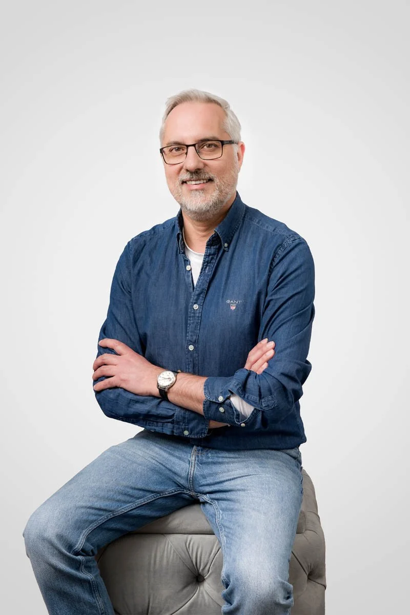 Homem de cabelo grisalho, barba e óculos, sentado em um banco cinza, usando uma camisa jeans e calça jeans, sorrindo com os braços cruzados, contra fundo branco.