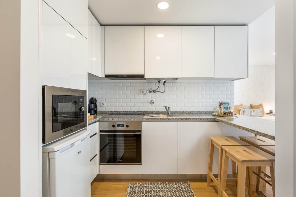 Cozinha moderna com armários brancos, eletrodomésticos embutidos, bancada de granito, paredes de azulejos brancos, duas banquetas de madeira ao lado da bancada.