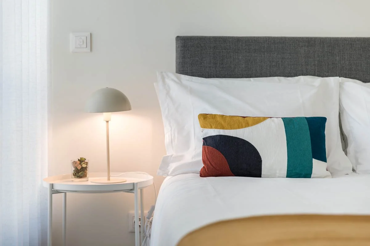 Cama com travesseiro colorido e cabeceira cinza, luminária de mesa e jarra na mesinha ao lado, parede branca