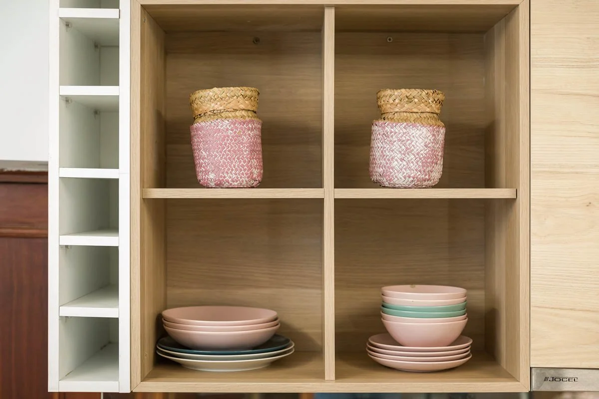 Estante de madeira com duas seções. No topo de cada seção, há uma cesta decorativa de fibra natural, uma com detalhes rosa e a outra com detalhes rosa e branco. Na seção esquerda, há tigelas cor de rosa empilhadas, e na seção direita, tigelas de dife