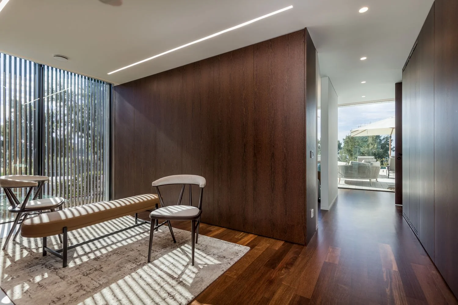 Sala de estar moderna com piso de madeira, parede de madeira escura e portas de vidro com cortinas verticais. Há uma cadeira branca e um banco de canto, com uma área de descanso ao ar livre com sofá, mesa e guarda-sóis ao fundo.