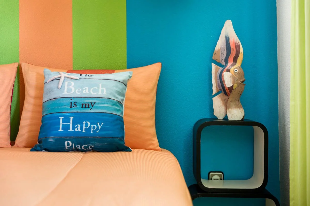 Cama com travesseiro decorativo com a frase "The Beach is my Happy Place", parede colorida com listras verdes, laranja e azul, quadro decorativo de uma tartaruga na parede azul, mesa preta ao lado da cama, cortina verde ao lado