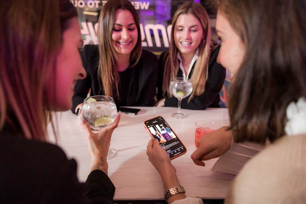 Quatro mulheres jovens conversando em um bar, uma delas segurando um drinque e visualizando vídeos no celular