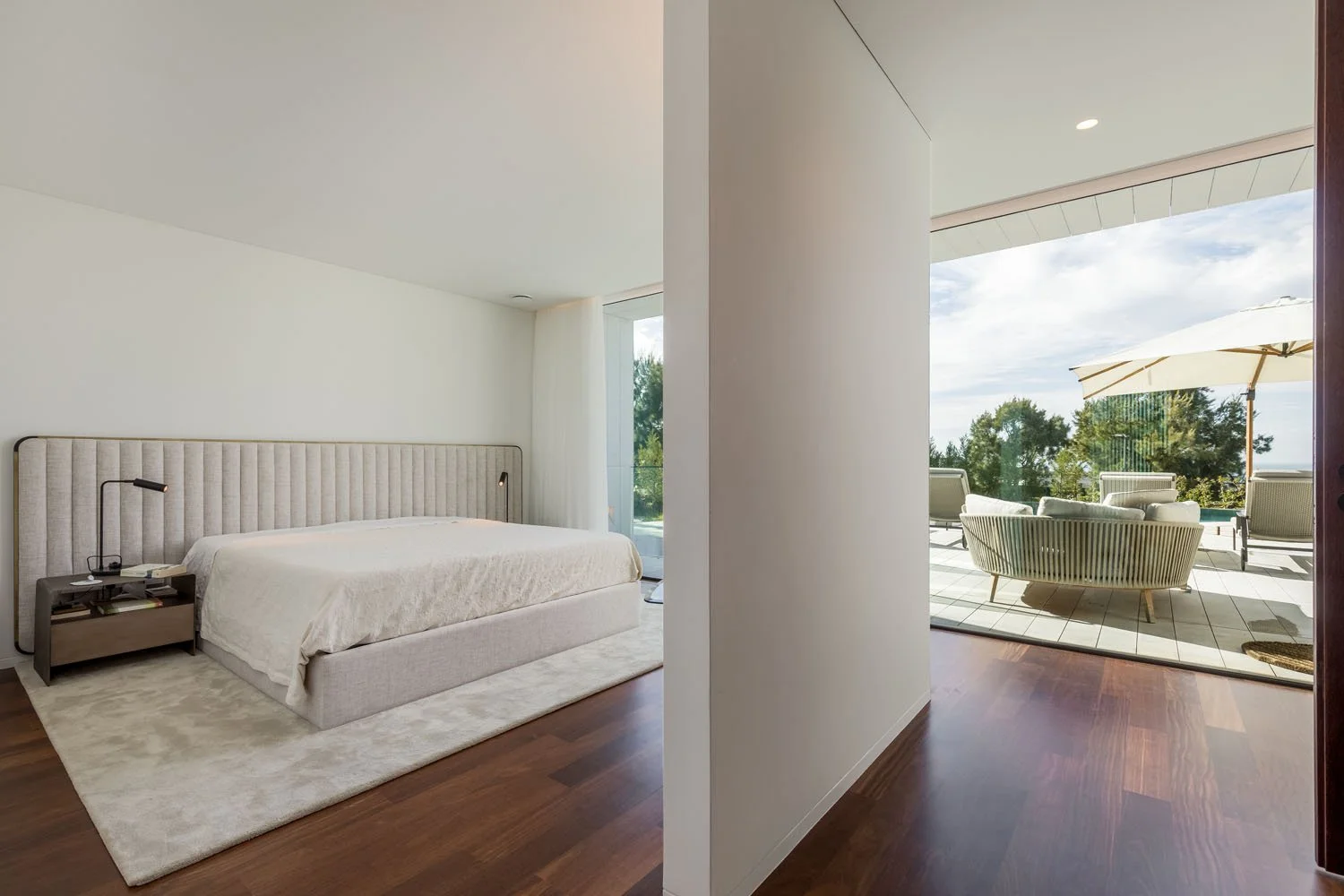 Quarto de design minimalista com cama grande, cabeceira almofadada, luminária de leitura ao lado, tapete claro e parede branca, com acesso a uma varanda com mobiliário externo e guarda-sol.