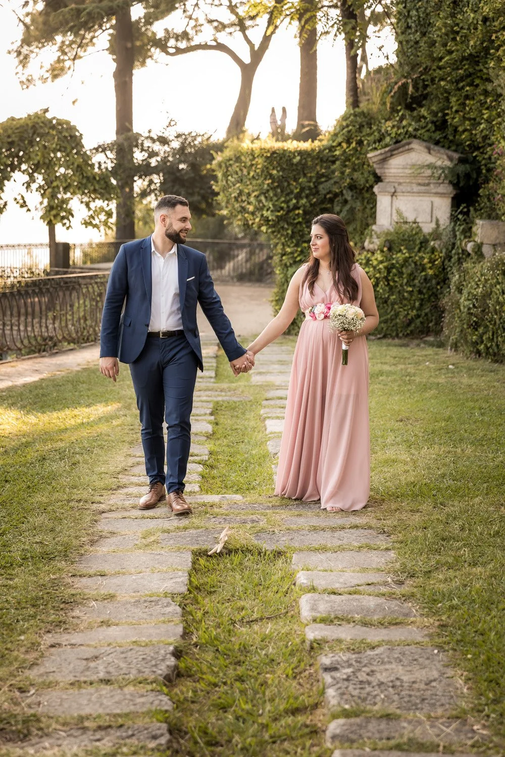 Casal caminhando de mãos dadas em um jardim ao pôr do sol, com ela segurando um buquê de flores e vestida com um vestido rosa longo, ele usando um terno azul escuro.