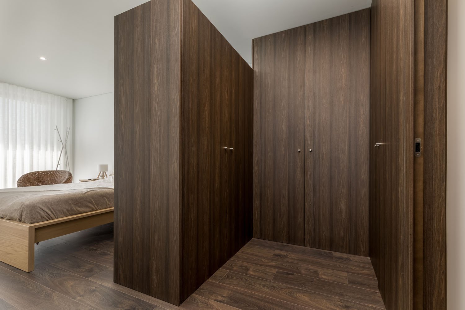 Closet de madeira escura ao lado de um quarto com cama, cortinas brancas e cadeira de vime.