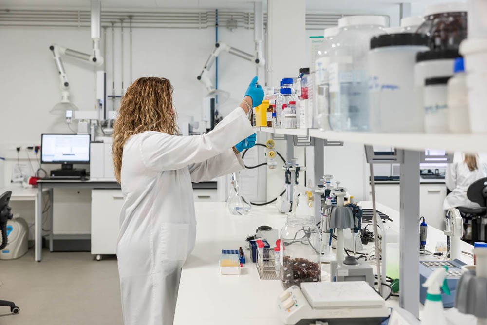 Pessoa realizando experimento em laboratório de pesquisa científica, manipulando líquidos e instrumentos em bancada de trabalho com equipamentos laboratoriais ao fundo.