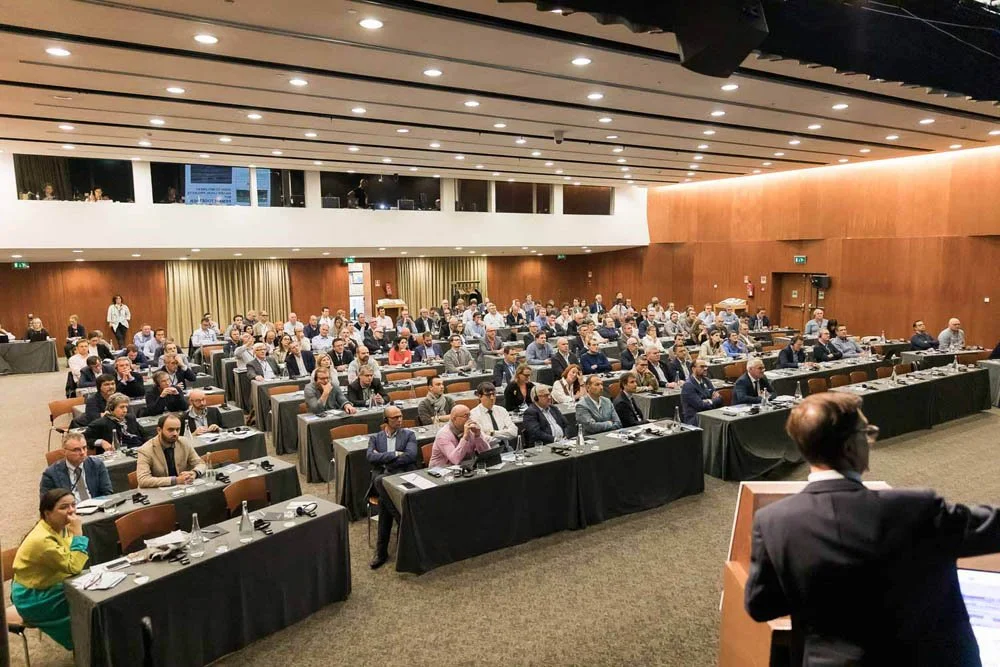 Uma sala de conferências lotada com várias fileiras de mesas e cadeiras, pessoas atendendo a uma apresentação. No palco, uma pessoa de terno fala para o público.