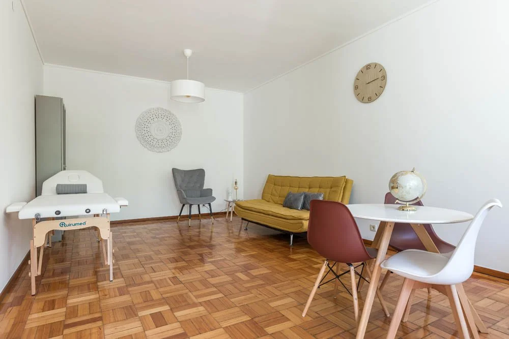 Sala de consultas com mesa de exame, sofá amarelo, cadeiras, relógio na parede, globo terrestre na mesa, piso de parquet.