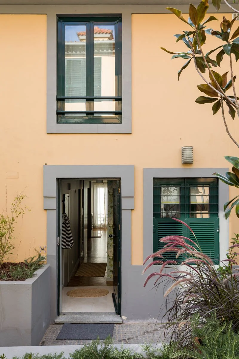 Fachada de uma casa com portas e janelas de moldura cinza, parede amarelo claro, vegetação e plantas ao redor, e jardim na entrada.