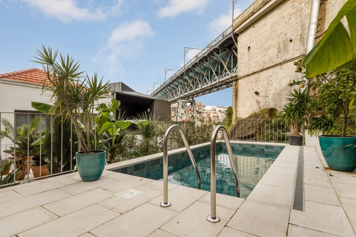 Piscina pequena com azulejos azuis, cercada por plantas em vasos e uma cerca branca, com uma ponte ou estrutura metálica suspensa ao fundo e céu claro.