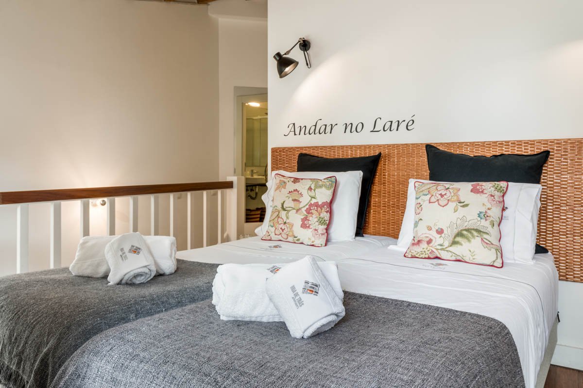 Cama de hotel com travesseiros e toalhas dobradas, parede com frase "Andar no Laré" e iluminação ambiental.