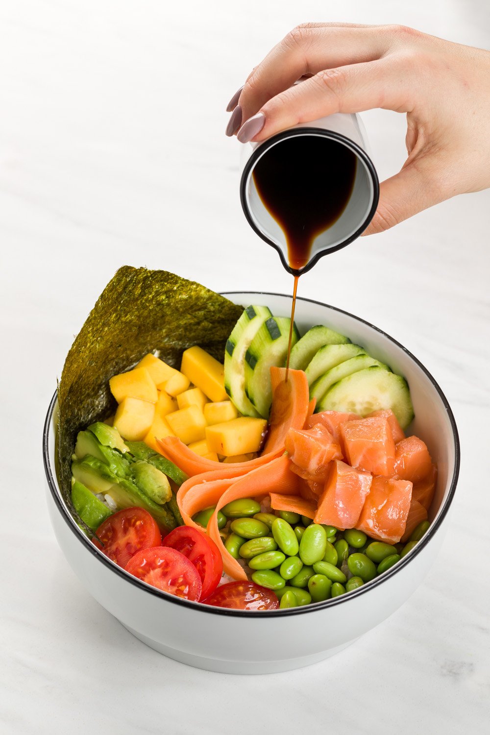 Pote com salmão, tomate, abacate, pepino, edamame, alface, salsicha de salmão, gergelim, alga e molho shoyu sendo despejado.