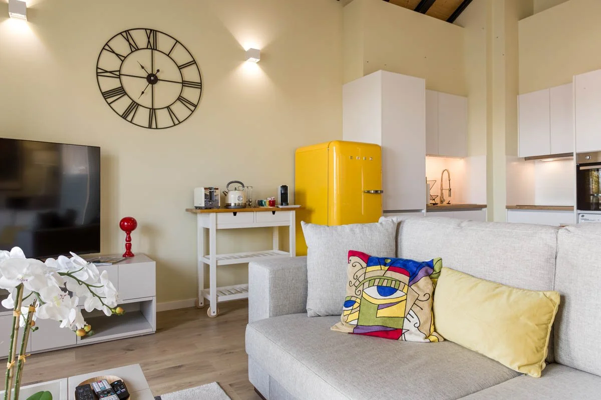 Sala de estar com sofá cinza, almofadas coloridas, televisão de tela plana, parede com relógio grande de ferro, mesa pequena com eletrodomésticos, geladeira amarelo, cozinha ao fundo com armários brancos e pia.