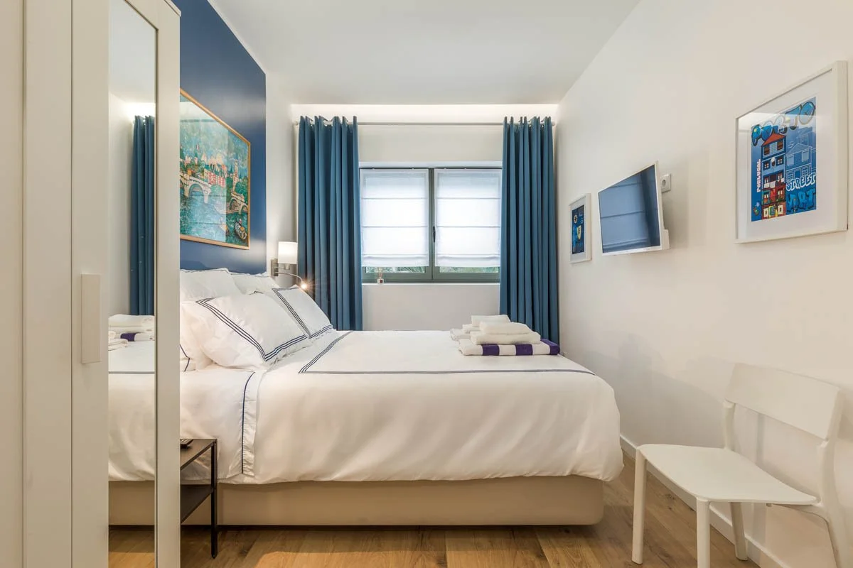 Quarto de hotel com cama de casal, cortinas azuis, quadro na parede e televisão.