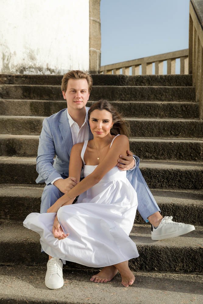 Casal sentados nos degraus de uma escada de pedra, com fundo de céu azul e uma estrutura de madeira. A mulher veste um vestido branco e o homem um terno cinza claro. Ela está descalça, ele com tênis branco.