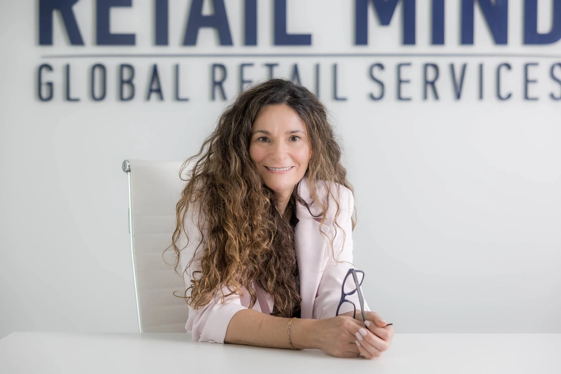 Mulher sorridente com cabelos cacheados longos sentada em uma mesa branca, usando blazer rosa claro, em um escritório com parede branca e placa com o texto 'RETAIL MIND' ao fundo.