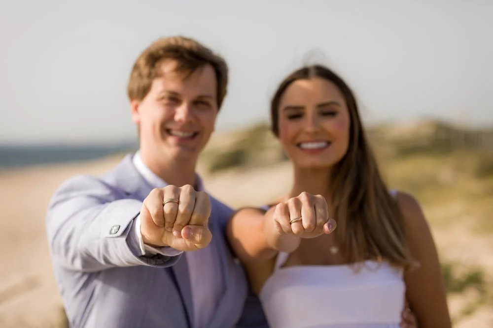 Casal feliz usando alianças de casamento na praia, mostrando seus anéis com sorriso.