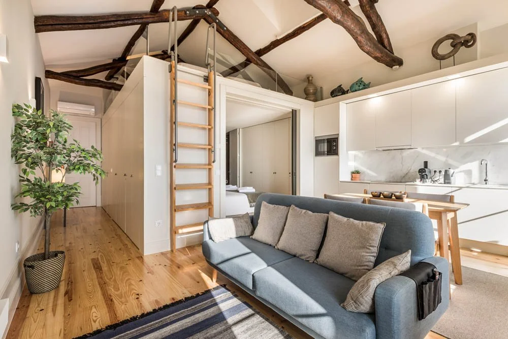 Sala de estar com sofá azul, almofadas cinza, cozinha de fundo com armários brancos, bancada de mármore, mesa de jantar de madeira, teto com vigas de madeira expostas, escada de peroba de pá, planta junto à parede.