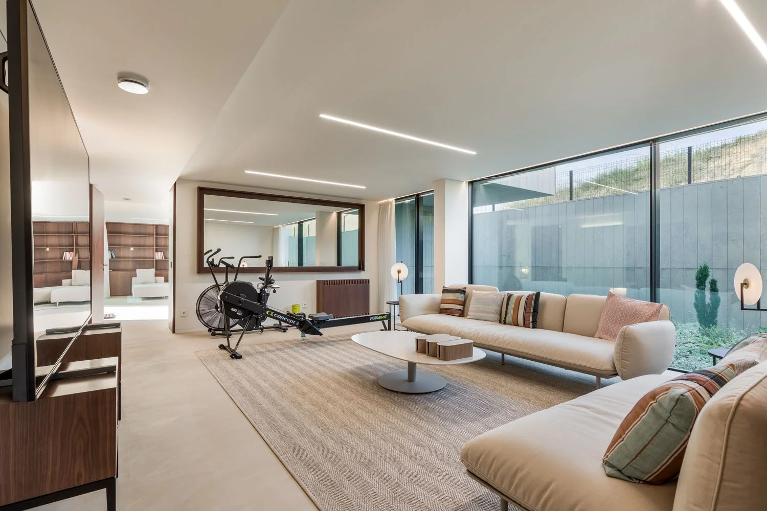 Sala de estar com sofás brancos, mesa de centro oval, tapete bege, uma bicicleta ergométrica ao fundo, grande janela de vidro com vista para o exterior.