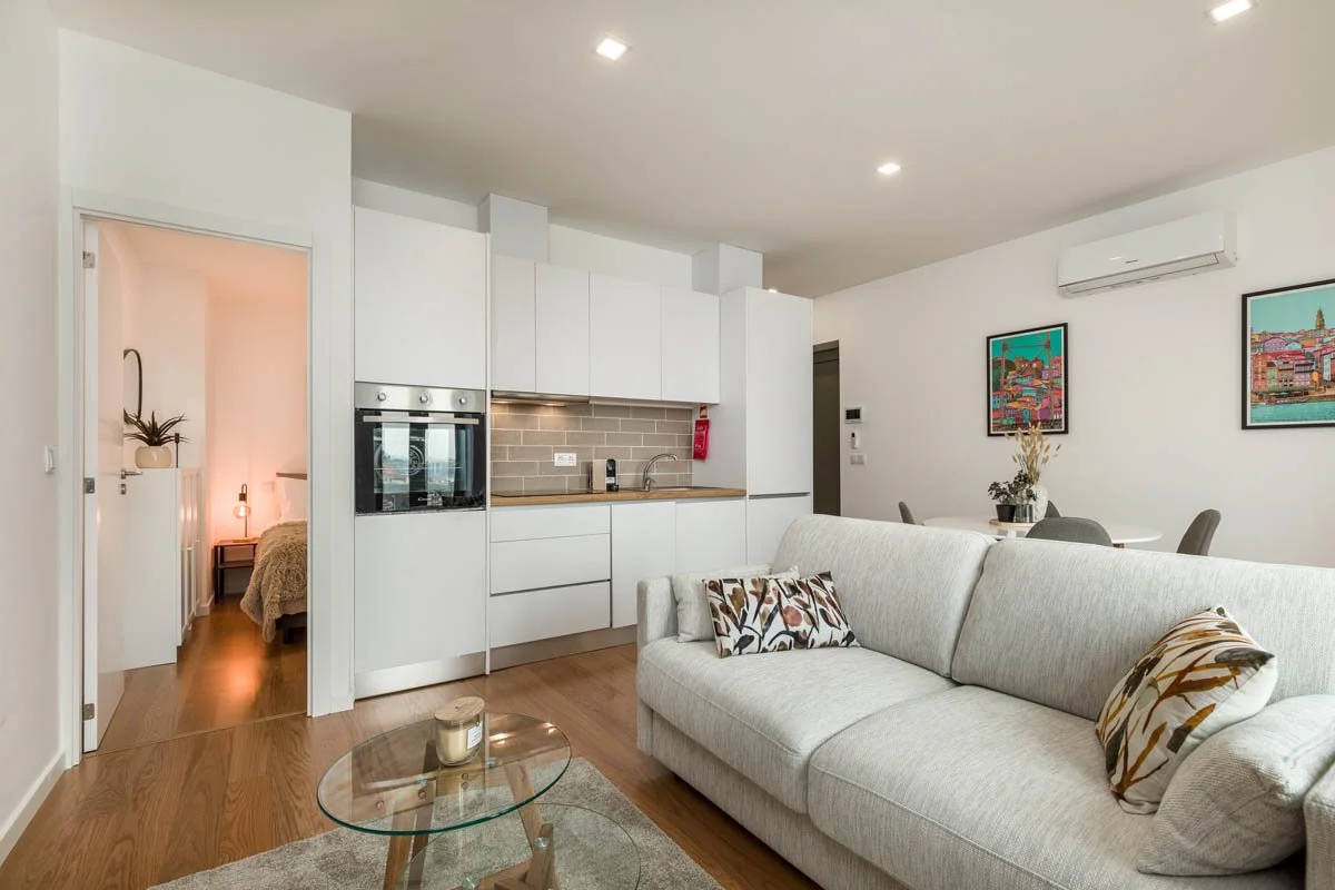 Sala de estar com sofá cinza claro, almofadas decorativas e mesa de vidro, cozinha branca com armários e eletrodomésticos, parede de azulejos cinza ao fundo, sala de jantar com mesa e cadeiras, paredes decoradas com quadros e ventilador de ar condici