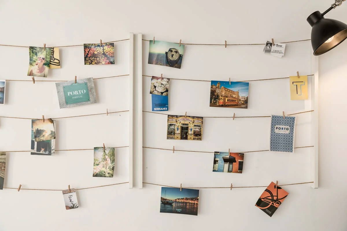 Parede branca com quatro linhas de cordão, cada uma presa com pregadores de roupas, exibindo várias fotos e cartões. Há uma luminária preta direcionada para a parede no canto superior direito.