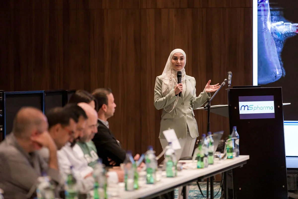 Mulher vestida formalmente, usando hijab, falando em um microfone durante uma apresentação em um painel de conferência, com várias pessoas sentadas na mesa à sua frente.