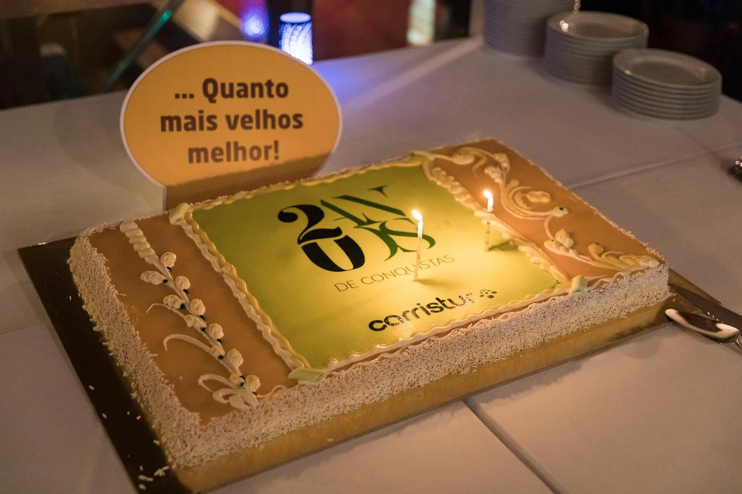 Bolo comemorativo do aniversário de 20 anos da Carriust, com velas acesas e mensagem, em comemoração aos 20 anos de conquistas da empresa, com uma placa ao lado que diz 'Quanto mais velhos melhor!'.