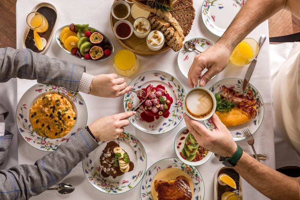 Mesa posta com diversos alimentos de café da manhã, incluindo frutas, bolo, waffle, ovos mexidos, sucos, café, pão e bacon, com várias mãos se servindo.