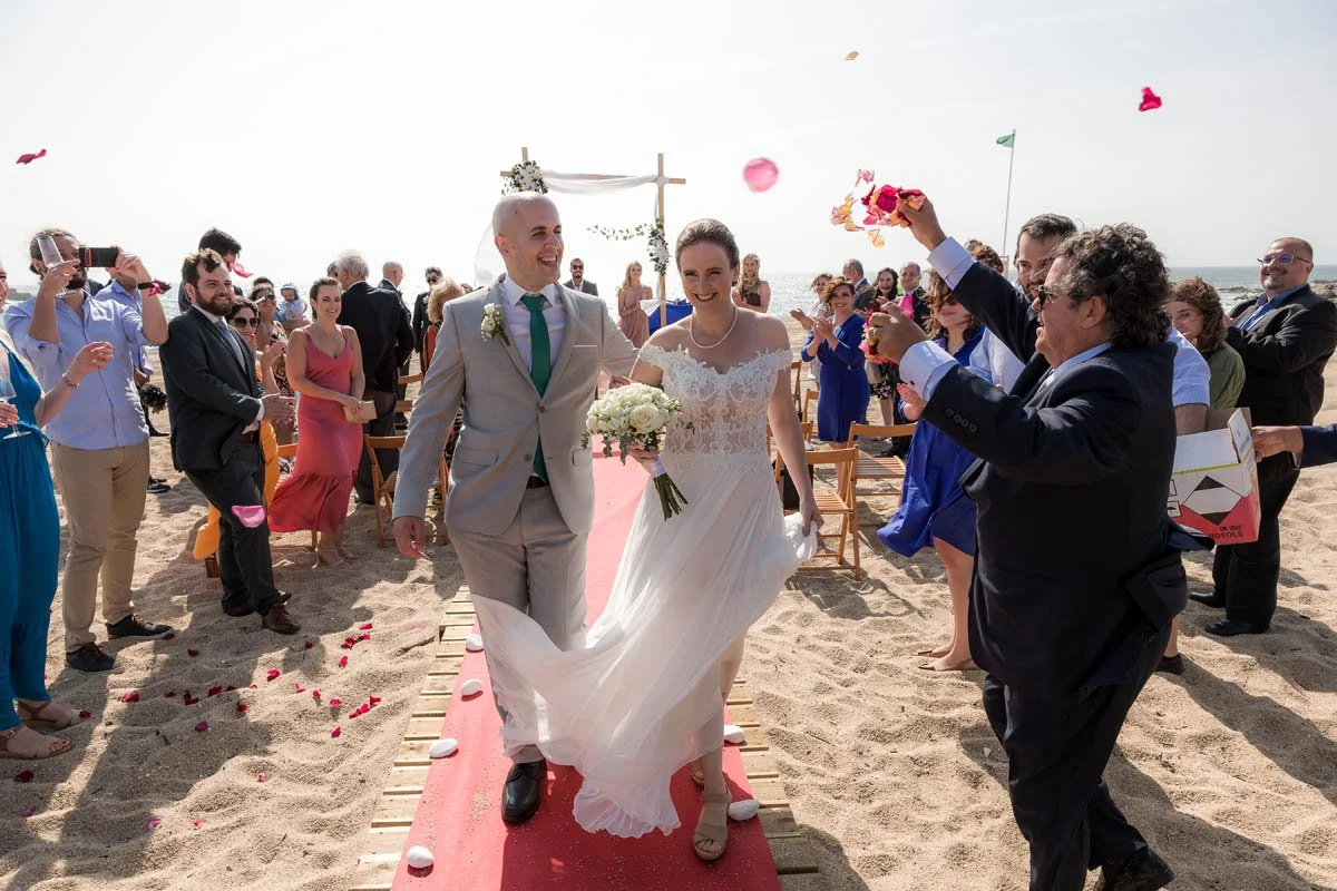 Casal de noivos caminhando pelo tapete vermelho na praia, rodeados por amigos e familiares celebrando o casamento ao ar livre, com o mar ao fundo.