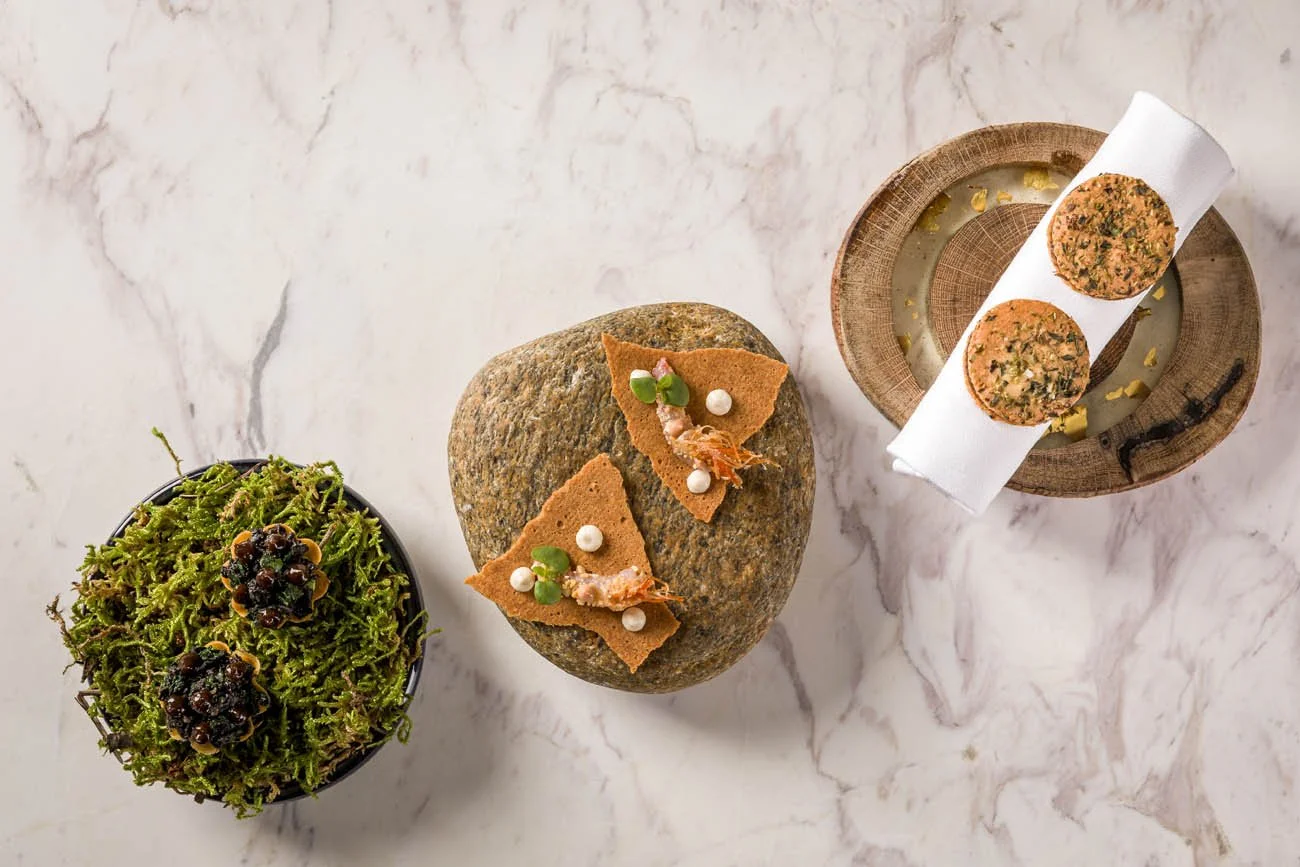 Prato de comida com três tipos de aperitivos em diferentes suportes, incluindo uma salada de ervas, uma pedra com aperitivos pequenos e um prato com biscoitos de avena.