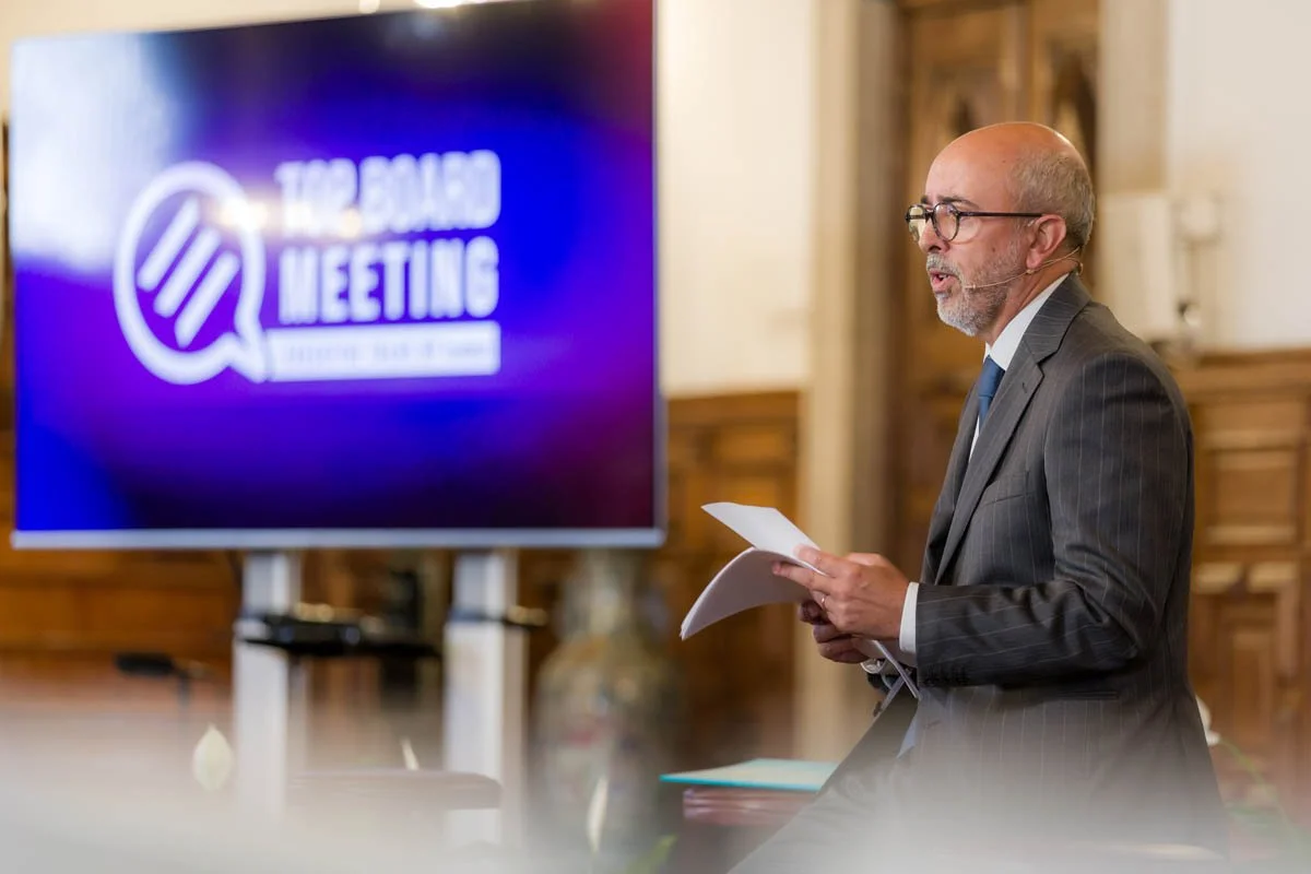 Homem de terno apresentando em uma reunião com tela de fundo que diz 'The Board Meeting'.